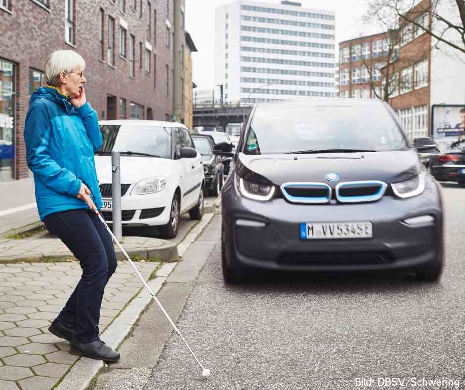 Farbfoto: Eine grauhaarige Frau mittleren Alters mit blauer Jacke, dunkler Hose und einem Langstock betritt eine Straße, auf der sich ihr ein dunkles Auto nähert. Ihr Gesichtsausdruck ist erschrocken, ihre linke Hand liegt am Gesicht.