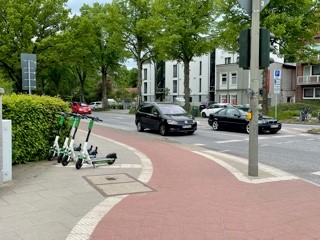 Eine Reihe E-Roller sind auf dem schmalen Gehweg aufgereiht. Wenn man diesen ausweichen möchte, steht man sofort auf dem radweg. An einer Stelle, die durch eine Kurve von Radfahrern schlecht überblickt werden kann.