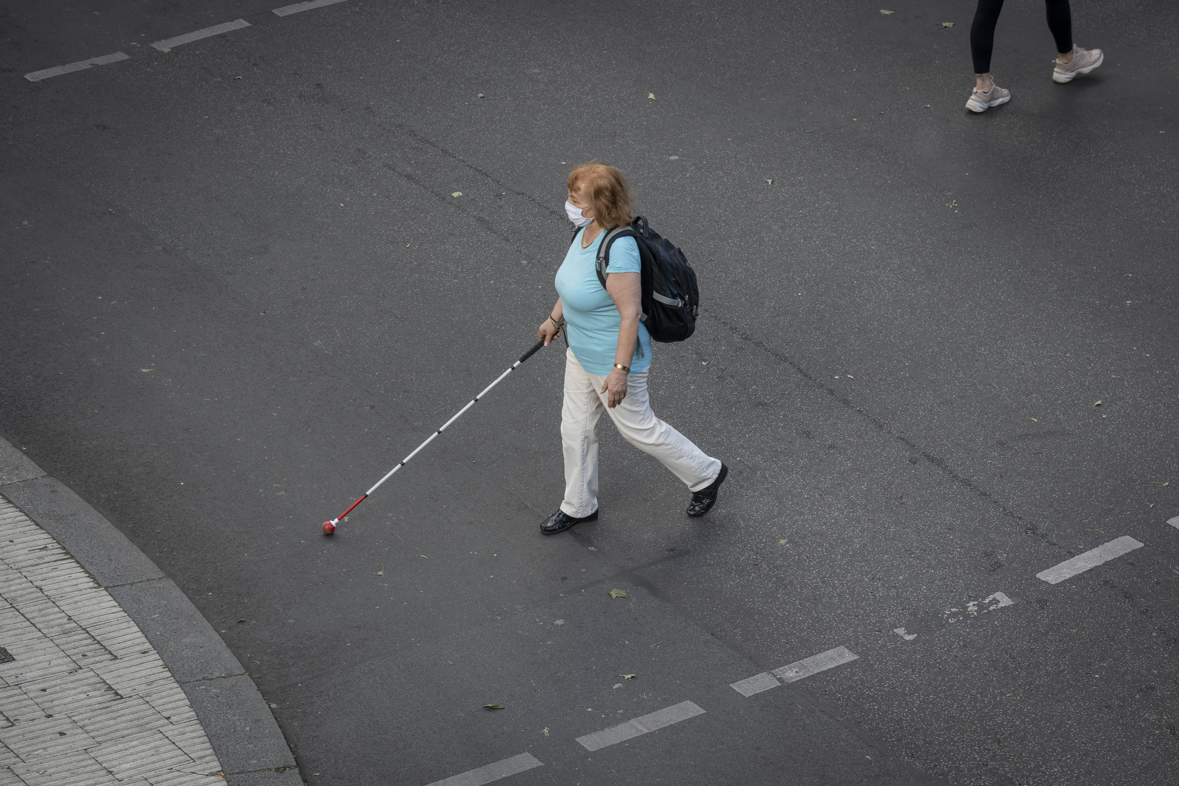 Eine Frau mit Mund-Nasen-Schutz überquert die Straße. Sie ist blind und orientiert sich mit Langstock